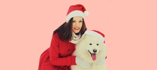 Christmas portrait of happy woman with white Samoyed dog in red santa hat on pink background