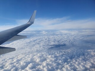 Airplane wing over clouds and sunny blue sky. Aerial view from an aircraft of an overcast sky with cumulus until the horizon and the sea appears through a hole on the stratocumulus.