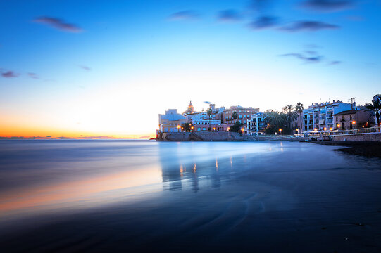 Fotografia de larga exposici&oacute;n de Sitges desde la playa San Sebasti&agrave; con la igl&eacute;sia de Sant bartomeu iluminada al fondo y un cielo con nubes al atardecer