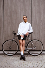 Stylish young african american woman holding sports bottle and looking at camera near bicycle on street 