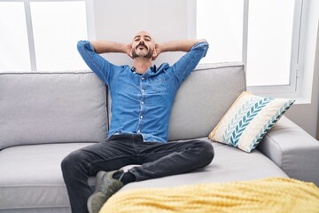 Young hispanic man relaxed with hands on head sitting on sofa at home