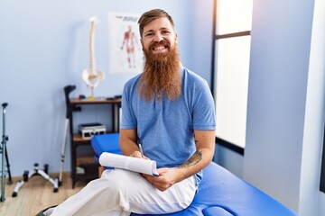 Young redhead man wearing physiotherapist uniform writing medical report at physiotherapy clinic