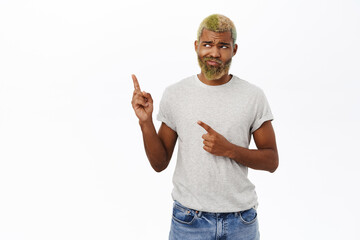 Naklejka premium Upset hipster guy with yellow beard and hair, points left, frowns and looks sad, stands over white background