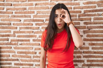 Young teenager girl standing over bricks wall doing ok gesture shocked with surprised face, eye looking through fingers. unbelieving expression.