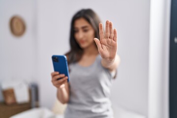 Young hispanic girl using smartphone doing stop gesture with hand at bedroom