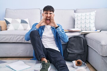 Young hispanic man sitting on the floor studying for university shouting angry out loud with hands over mouth