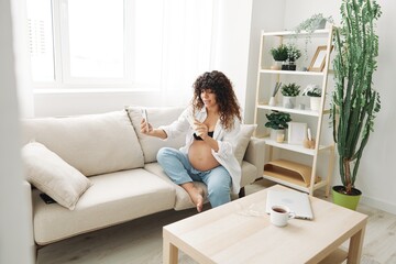 Pregnant woman smile blogger showing baby feeding bottle filming herself on phone, video chat sitting on sofa at home freelancer in last month of pregnancy