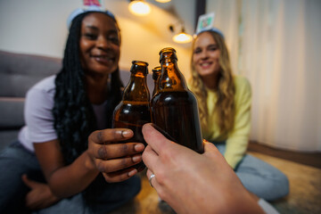 Girls looking at the camera, multiracial friends toasting with bear or cider sitting on a floor