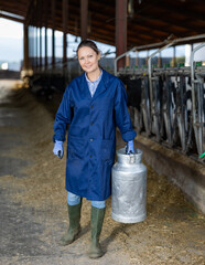 Female owner with milk can standing in stall on background with herd of cows on farm