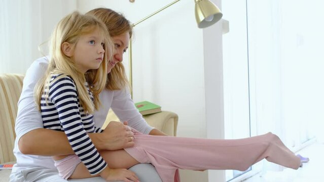 Caring loving mother dressing her daughter by the window.