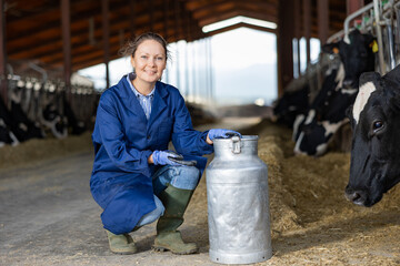 Positive woman farmer squatting at milk can in cowshed and smiling.
