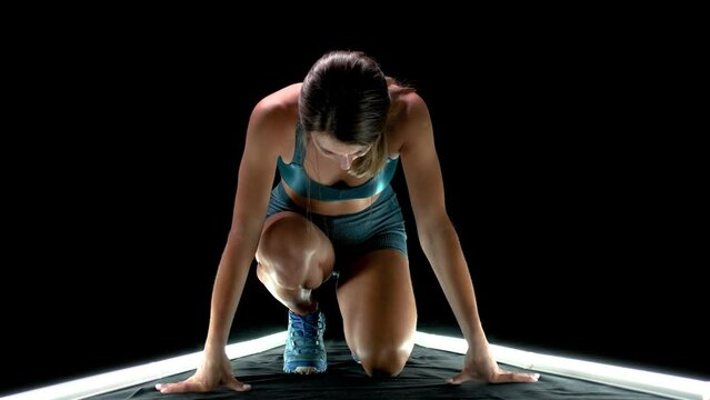 Female runner in race  start position. Girl in sportswear posing on lit track against dark background.