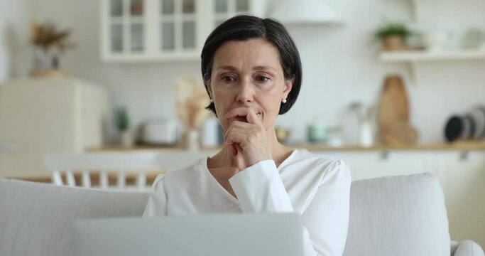 Serious middle-aged woman working on laptop at home looks at device screen thinks, search problem solutions feels undecided or hesitated, text email to client do telework remotely using internet. Tech