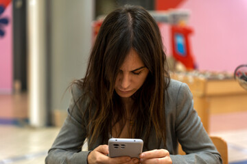 Concentrated young female entrepreneur with long dark hair in formal clothes messaging on mobile phone while sitting in modern cafe