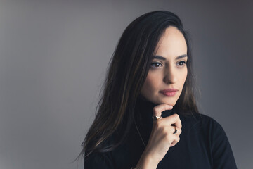 Young latin american woman with long dark hair dressed in black turtleneck looking into distance with pensive expression and holding finger to chin. Grey background horizontal studio shot. High