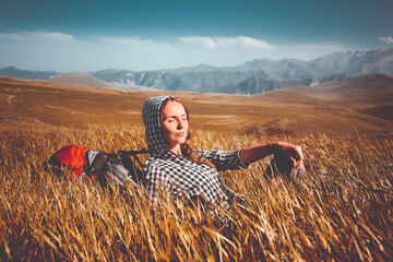 Tourist woman relax sitting in orange grass field. Mountain range in background. Autumn colorful scene. Backpack girl enjoy sunset light. Stunning nature scenery. Travel, adventure, concept image