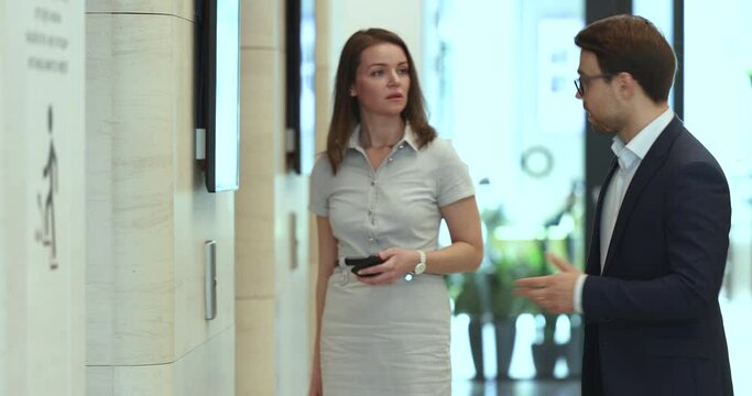 Two colleagues, businessman and businesswoman having conversation, walking in office hallway, calls the elevator, discuss collaborative project or common task during working day in modern workspace