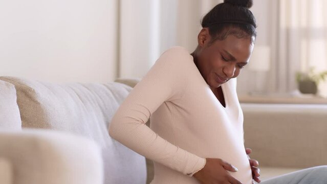 Training contractions. Young worried pregnant black woman feeling pain attack in stomach, deeply breathing at home
