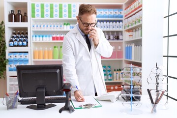 Middle age man pharmacist talking on telephone writing on document at pharmacy