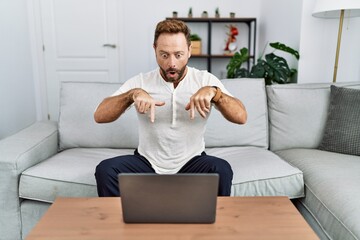 Middle age man using laptop at home pointing down with fingers showing advertisement, surprised face and open mouth