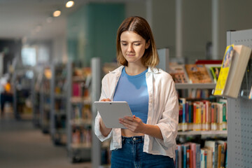 Young beautiful woman using digital tablet, searching online. Female student in library, education...