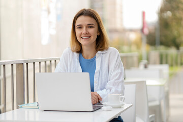 Fototapeta na wymiar Young smiling caucasian woman using laptop computer looking at camera sitting in cafe terrace. Portrait of happy successful freelancer at workplace 