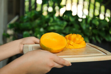 Woman holding wooden board with cut ripe mango outdoors, closeup