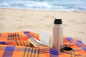 Metallic thermos with hot drink, open book and plaid on sandy beach near sea