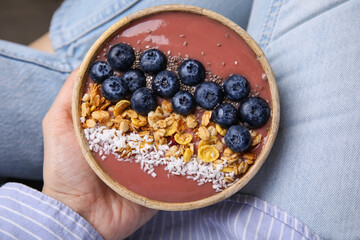 Woman holding bowl of delicious fruit smoothie with fresh blueberries, granola and coconut flakes, top view