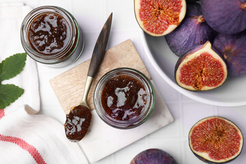 Glass jars of tasty sweet fig jam and fruits on white tiled table, flat lay