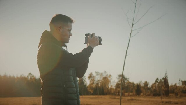 Male photographer alone in open field out in nature at sunset. Lens flare caused by sunlight. Caucasian young man taking landscape photos with dslr camera. Travel blogger taking pictures. Slow motion.