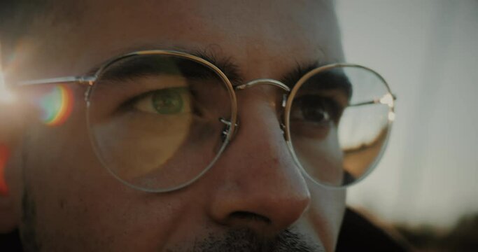 Eyes close-up of an angry and serious Caucasian young man with glasses having difficulty seeing. Generation z lifestyle. Portrait of a thoughtful and focused millennial blinking his eyes at sunset.