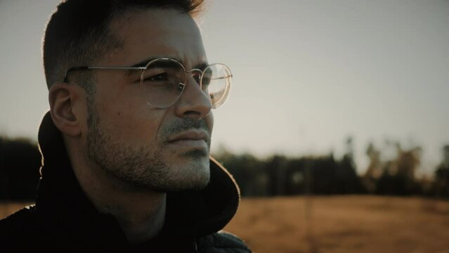 Face close-up shot of a young and handsome Caucasian man wearing glasses. Unhappy male outdoors thinking of problems, struggling with mental health, depression, and sadness. Side view. Slow motion.