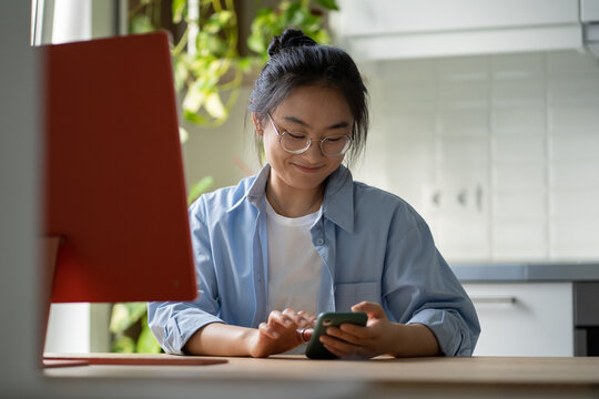 Smiling Korean Girl Remote Employee Sitting At Home Office Using Cellphone For Personal During Work Hours, Chatting On Mobile Phone While Working Remotely. Procrastination And Freelance Career