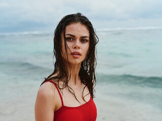 Beautiful sexy woman in a red swimsuit looks at the camera on the ocean beach with wet hair