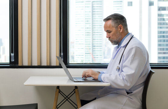 Senior professor with mustache and beard typing on laptop computer keyboard. Sitting near the window in cafeteria. Outside the window is a city view with high rise buildings.