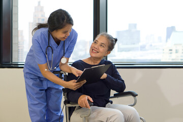 Woman physical therapist in blue uniform with stethoscope and clipboard explaining rehabilitation steps to an elderly in a wheelchair.