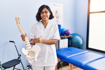 Young latin woman wearing physiotherapist uniform holding anatomical model of vertebral column at...