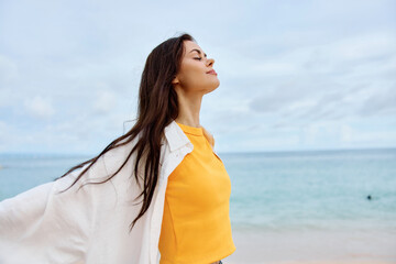 Happy woman smile with teeth with long hair brunette walks along the beach in a yellow t-shirt denim shorts and a white shirt near the sea summer journey and feeling of freedom, balance