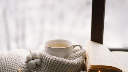 Cozy winter still life. Cup of hot tea and an open book with a warm sweater on a vintage wooden windowsill. Cozy home concept. Sweet home.