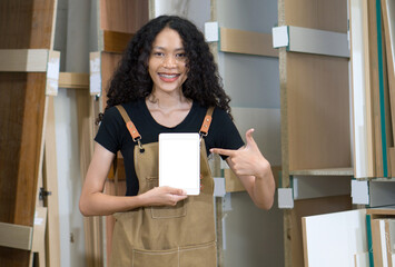 Teenager in brown apron pointing finger at white screen tablet computer. Plywood storage compartment are on the background. Atmosphere in a wooden furniture factory.