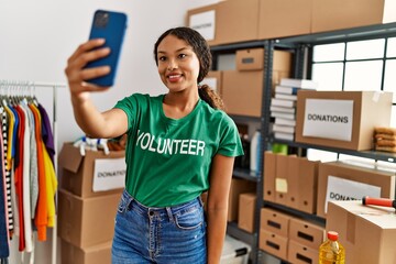 Young latin woman wearing volunteer uniform make selfie by the smartphone at charity center