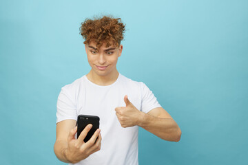 a happy man is standing on a light blue background and looking into his smartphone is talking via video link showing a thumbs up. Horizontal studio photography with empty space