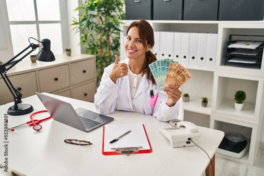 Sticker Hispanic doctor woman holding brazilian reals banknotes smiling happy and positive, thumb up doing excellent and approval sign