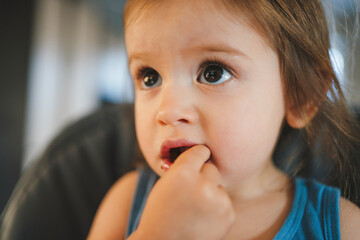 Close-up portrait of a little girl holding her fingers in her mouth, biting the snack she is having at the table. First meal, self-feeding