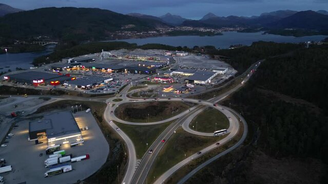 Drone flies slowly backwards over Road junction at Digernes, Skodje in &Aring;lesund municipality. Evening, with light from street lights and cars. View towards Skodje in the east and industrial area.