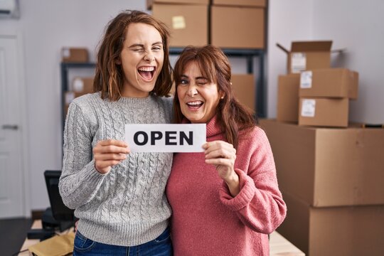 Two Women Working At Small Business Ecommerce Holding Open Banner Winking Looking At The Camera With Sexy Expression, Cheerful And Happy Face.