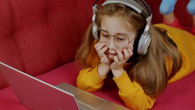 A cute teenage girl in a yellow sweatshirt, wearing large round glasses, lies on a dewan in front of a laptop with her chin in her hands and dangles her legs. Teens digital addiction problem concept