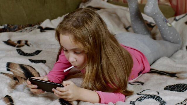 A cute blonde-haired girl lies on the sofa, her legs dangling, carefully looks at the smartphone screen and sucks a sweet lollipop. The concept of digital addiction in children and adolescents