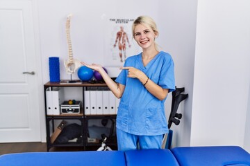 Young physiotherapist woman working at pain recovery clinic amazed and smiling to the camera while presenting with hand and pointing with finger.